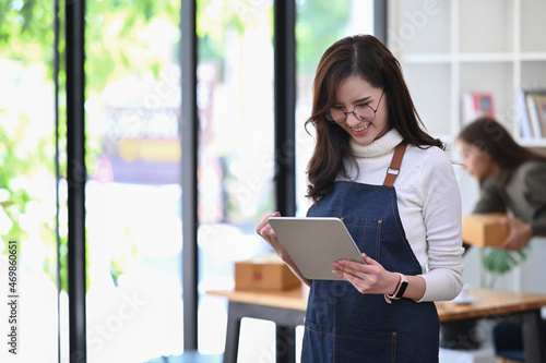 Billede på lærred Cheerful young Asian woman entrepreneur using digital tablet and standing in her coffee shop