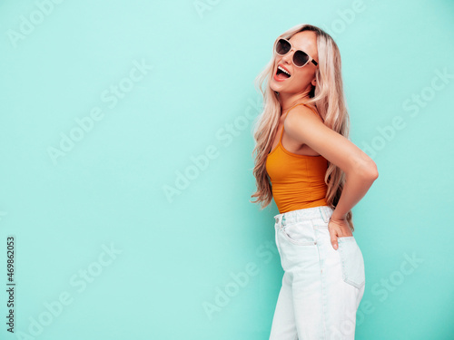 Portrait of young beautiful smiling female in trendy summer clothes. Sexy carefree blond woman posing near blue wall in studio. Positive model having fun indoors. Cheerful and happy. In sunglasses