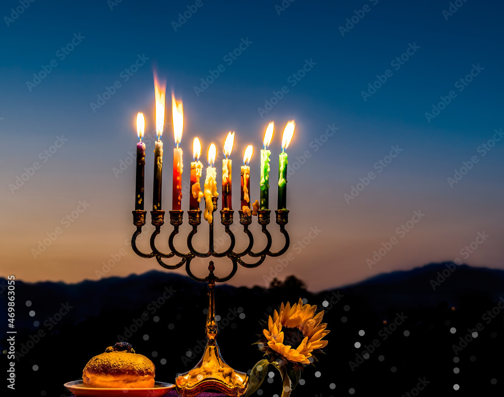 Fototapeta premium Close-up of festive burning wax candles that are traditional symbols of Hanukkah Holiday of Light, selective focus on burning candles and sunflower, blurred background with night mountains and sky 