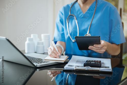 Pharmacist writing making notes in drug information using laptop computer sitting at desk. Woman physician, nurse or pharmacist wearing white coat writing in paper notebook.