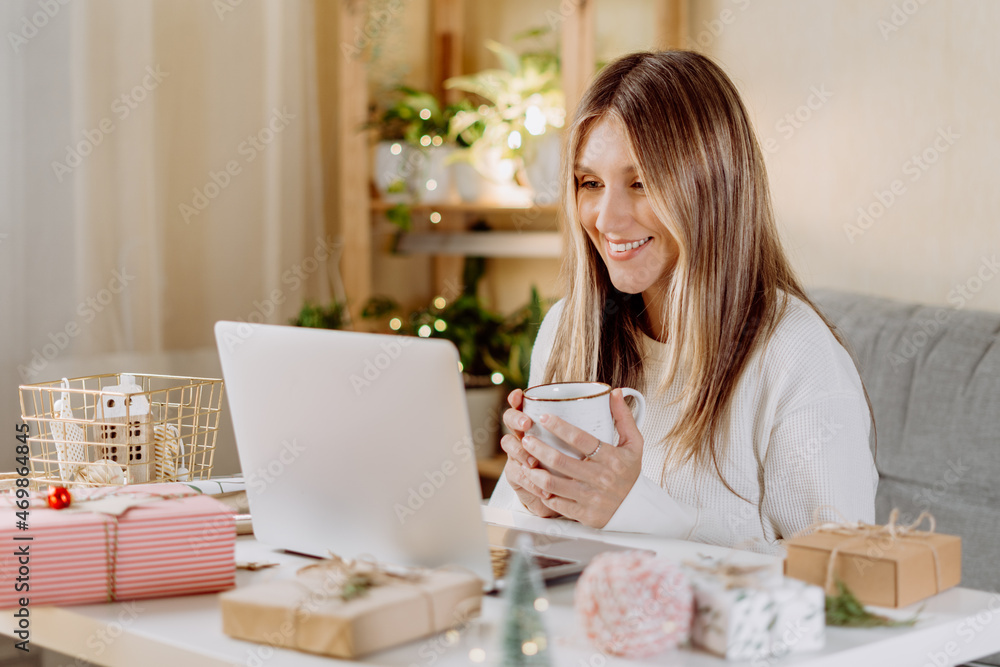 Happy and smiling young woman wrapping and preparing Christmas gifts, typing at laptop and having online video chat. Online shopping at Christmas holidays. Home interior with lights and plants Happy and smiling young woman wrapping and preparing Christmas gifts, typing at laptop and having online video chat. Online shopping at Christmas holidays. Home interior with lights and plants