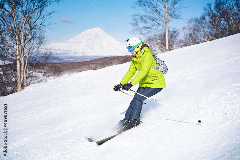 Cheerful skier girl in green jacket in front of snowy mountains and blue sky Cheerful skier girl in green jacket in front of snowy mountains and blue sky