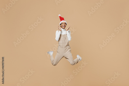 Full body surprised young african man 20s wear Santa Claus red Christmas hat holding face jump high isolated on plain pastel beige background studio portrait. Happy New Year 2022 celebration concept. © ViDi Studio