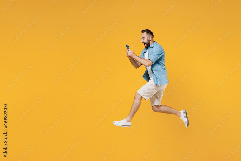 Full body side view young happy caucasian man 20s wearing blue shirt white t-shirt jump high use hold mobile cell phone isolated on plain yellow background studio portrait. People lifestyle concept.