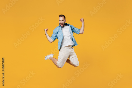 Full body young overjoyed satisfied happy caucasian man 20s wearing blue shirt do winner gesture clench fist jump high isolated on plain yellow background studio portrait. People lifestyle concept