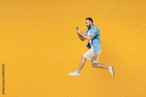 Full body side view young happy caucasian man 20s wearing blue shirt white t-shirt jump high use hold mobile cell phone isolated on plain yellow background studio portrait. People lifestyle concept. © ViDi Studio