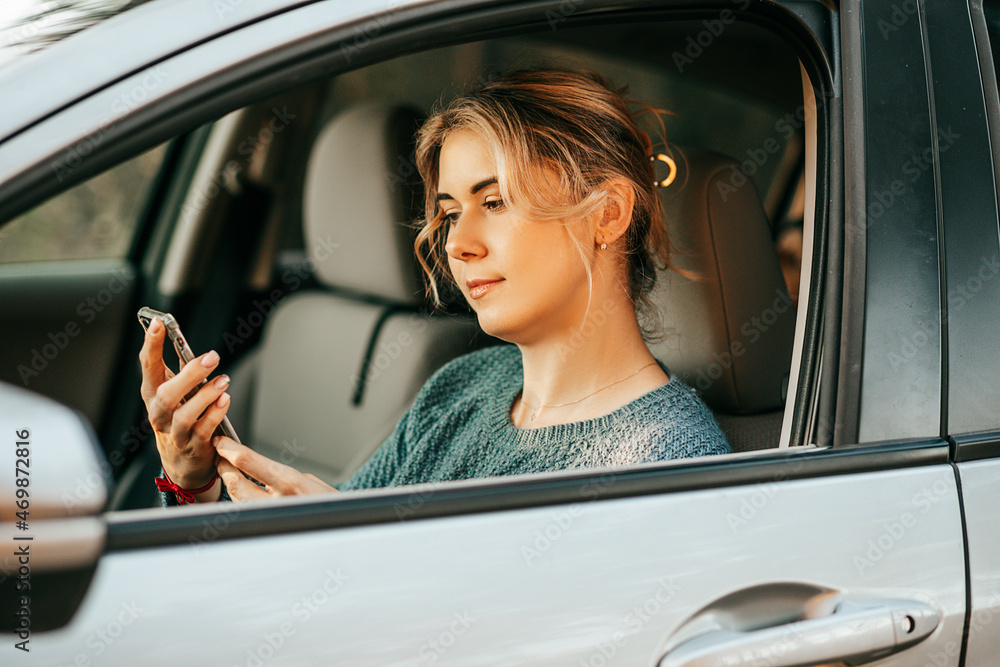 Young beautiful woman sits behind wheel of car, looks at screen of mobile phone, typing sms massage uses smartphone, navigation system, map, assistance in finding out