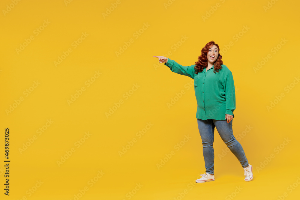 Full size body length excited happy vivid young ginger chubby overweight woman 20s wears green shirt go point on workspace area copy space mock up isolated on plain yellow background studio portrait.