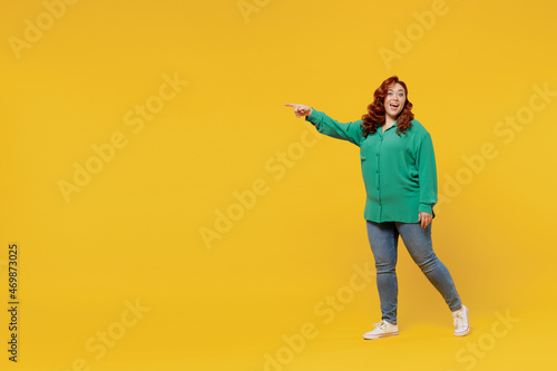 Full size body length excited happy vivid young ginger chubby overweight woman 20s wears green shirt go point on workspace area copy space mock up isolated on plain yellow background studio portrait.