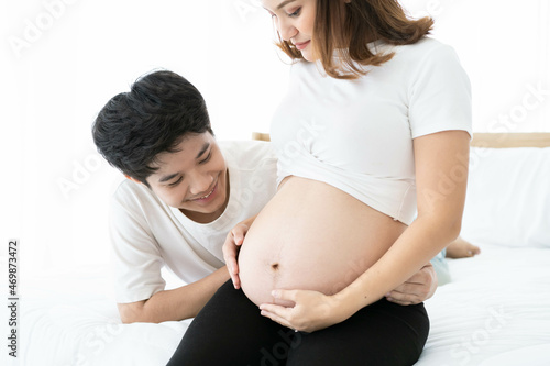 Lovely Asian husband gently looking at a belly of his pregnant wife while laying on the bed together. Father feels excited while listening on his wife's stomach. Concept of happy family.