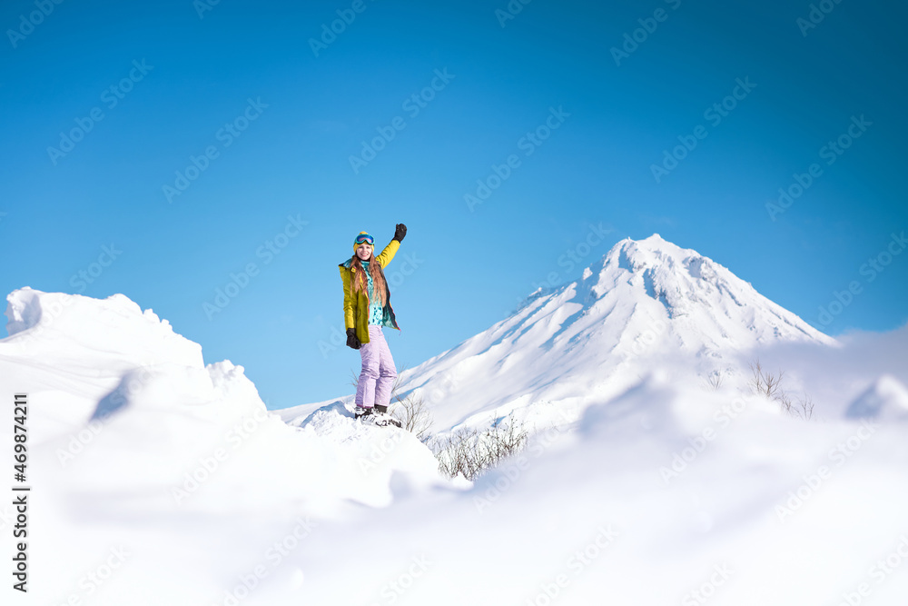 Cheerful girl snowboarder in yellow jacket in front of snowy mountains and blue sky