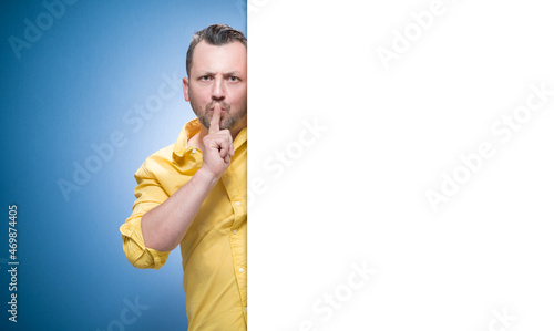 Hush. Man holding finger on lips, keep the secret concept over blue background, dresses in yellow shirt