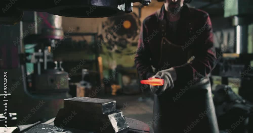 A blacksmith is creating a blank for an ax blade. He holds the hot metal with tongs and puts it under the press to flatten it A blacksmith is creating a blank for an ax blade. He holds the hot metal with tongs and puts it under the press to flatten it