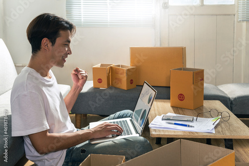 Cheerful Asian male freelance sits on a floor at home and works on a laptop computer having a new order on a monitor screen with the postal parcel for shipping. Succeed Online selling concept ideas