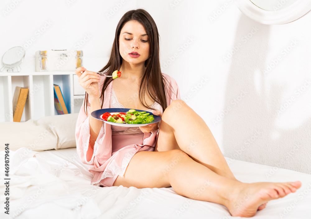 Positive pretty woman in bathrobe holding fork and eating vegetable salad in bed at home