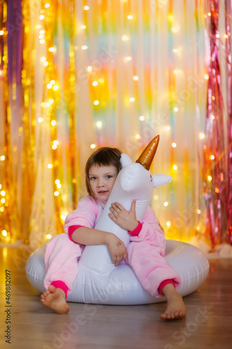 cute girl in a soft kigurumi unicorn costume playing with an inflatable ring unicorn on a colorful bright background, pajama theme party, rainbow theme and children emotions