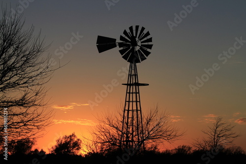A shot of a colorful Kansas Sunset with a farm Windmill silhouette with tree's and clouds north of Hutchinson Kansas USA out in the country in a farm pasture.