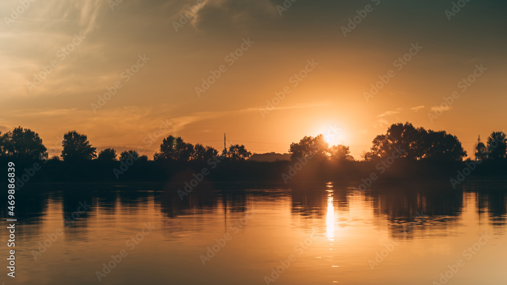 A warm orange sunset over the river. Trees and clouds reflected in the water. The ball of sun hides behind the forest. The beauty of nature.