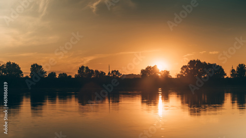 A warm orange sunset over the river. Trees and clouds reflected in the water. The ball of sun hides behind the forest. The beauty of nature.