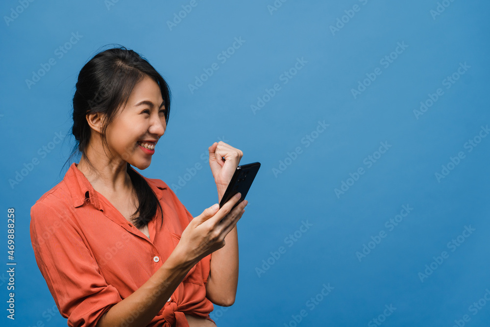 Surprised young Asia lady using mobile phone with positive expression, smiles broadly, dressed in casual clothing and stand isolated on blue background. Happy adorable glad woman rejoices success. Surprised young Asia lady using mobile phone with positive expression, smiles broadly, dressed in casual clothing and stand isolated on blue background. Happy adorable glad woman rejoices success.