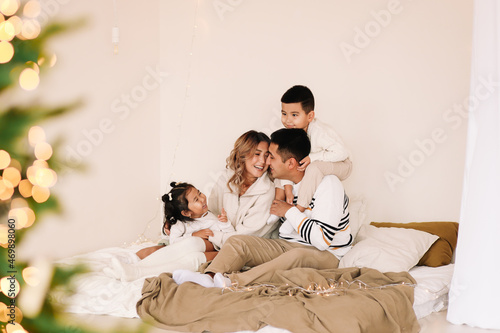 A mixed race Asian family with two children in cozy sweaters is relaxing together, resting and having fun on a bed in a decorated bedroom with Christmas tree at home in the New Year. Selective focus