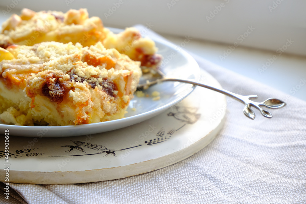 Two slices of homemade apple and apricot cheesecake pie with a beautiful stylish twig-shaped teaspoon on a wooden tray on a gray napkin on a white table. A healthy light lunch. Empty space. Two slices of homemade apple and apricot cheesecake pie with a beautiful stylish twig-shaped teaspoon on a wooden tray on a gray napkin on a white table. A healthy light lunch. Empty space.