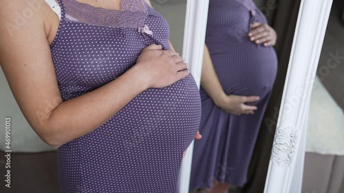 Woman waiting for child stands in a purple nightshirt in front of the mirror. Pregnant woman strokes her stomach with one hand reflecting in the mirror.