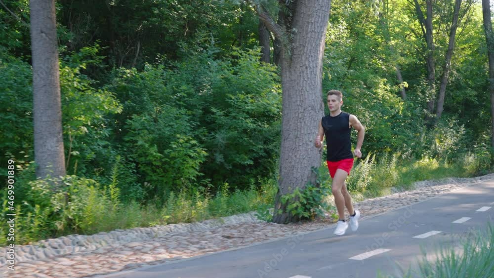Slow motion jogger in summer sportswear running on road with marking in bright sunshine, green trees on background. Tracking shot male athlete training in park in summer. Concept of fitness Slow motion jogger in summer sportswear running on road with marking in bright sunshine, green trees on background. Tracking shot male athlete training in park in summer. Concept of fitness