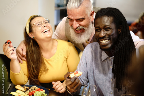Cheerful interracial group of adult friends eating, talking and joking together - multiracial group of adult people sitting at the dinner table having fun