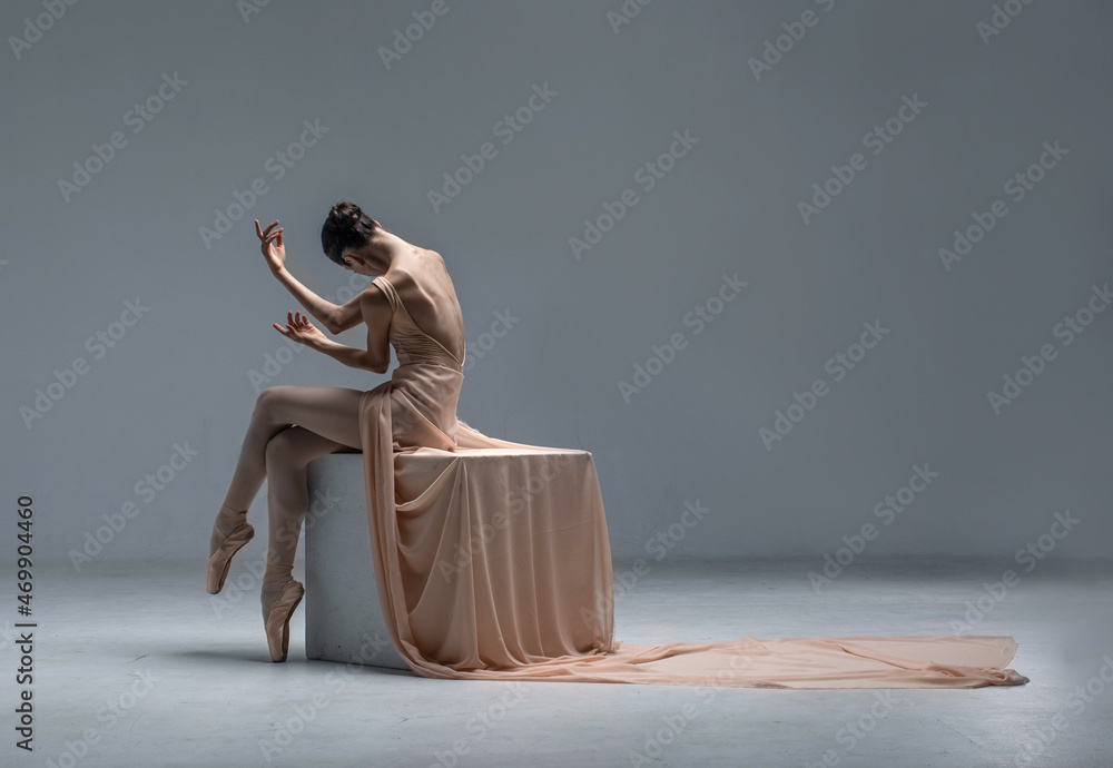 Beautiful ballerina sitting in white room on a white cube in in skin color leotard and long textile. She has pointe shoes on her feet Beautiful ballerina sitting in white room on a white cube in in skin color leotard and long textile. She has pointe shoes on her feet