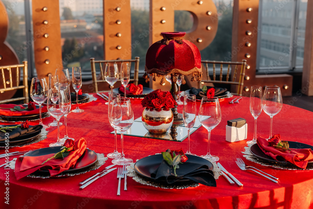 Guests table setting for banquet in black, red and gold style. Elegant  dinner: decor, tablecloth, plates with napkins and fresh roses, glasses,  cutlery. Themed party celebration on the roof, outdoor. Photos