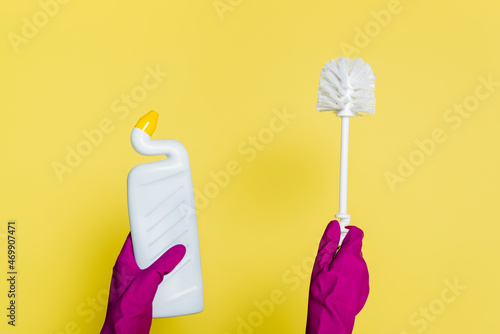 partial view of cleaner in rubber gloves holding bottle with detergent and toilet brush on yellow photo