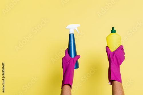 partial view of cleaner in rubber gloves holding detergent and spray bottle on yellow