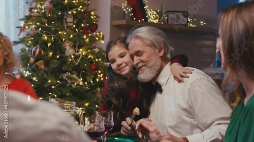 Happy caucasian grandfather sitting at the dinner table with his family celebrating Christmas. Little shy granddaughter giving huggs to grangfather while having Christmas dinner at home. photo