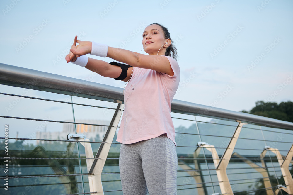 Beautiful woman, athlete wearing pink T-shirt stretches her body, standing on a glass city bridge during a morning workout at dawn. Sport, healthy and active lifestyle, vitality, weight loss concept Beautiful woman, athlete wearing pink T-shirt stretches her body, standing on a glass city bridge during a morning workout at dawn. Sport, healthy and active lifestyle, vitality, weight loss concept