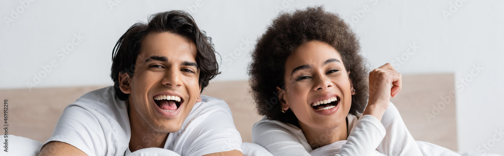happy multiethnic couple laughing while lying in bed, banner happy multiethnic couple laughing while lying in bed, banner