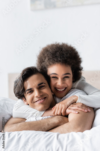 cheerful interracial couple smiling while hugging on bed