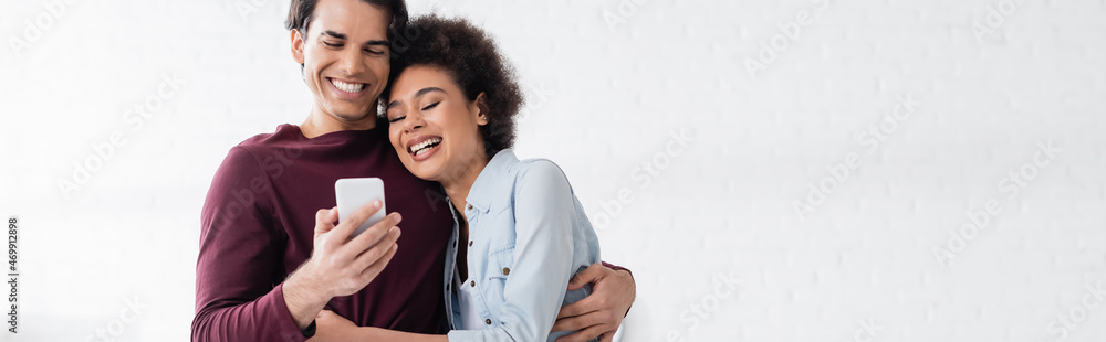 happy man using smartphone and hugging smiling african american girlfriend, banner happy man using smartphone and hugging smiling african american girlfriend, banner