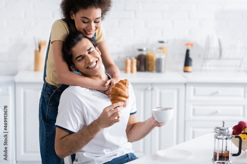 happy african american woman hugging boyfriend with cup of tea and croissant