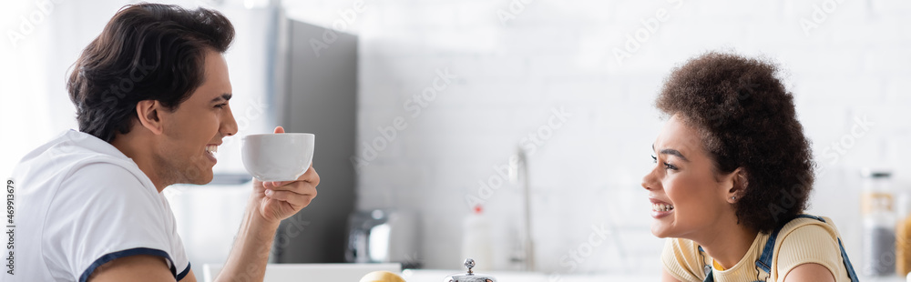 happy interracial couple smiling while drinking tea in morning, banner happy interracial couple smiling while drinking tea in morning, banner