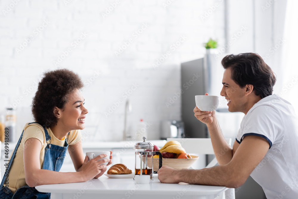 happy interracial couple smiling while looking at each other during breakfast