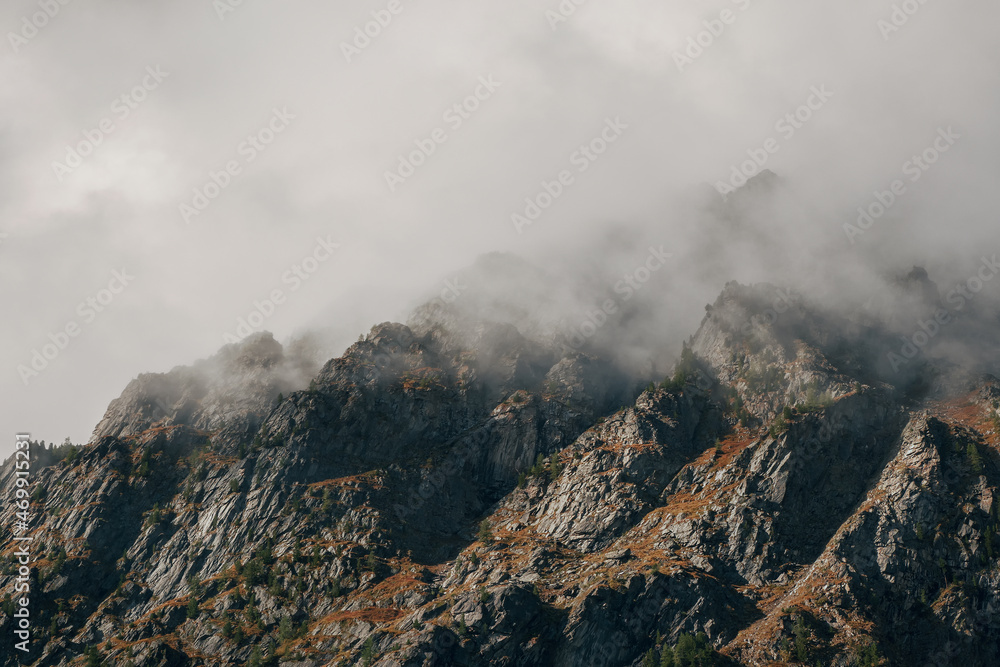 Dramatic fog among giant rocky mountains. Ghostly atmospheric view to big cliff in cloudy sky. Low clouds and beautiful rockies. Minimalist scenery mysterious place.