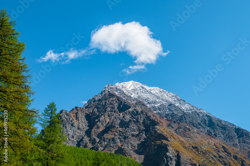 White clouds over a high pointed mountain. Snowy high-altitude alpine landscape with snow-capped mountain peak and sharp rocks under cloudy blue sky.