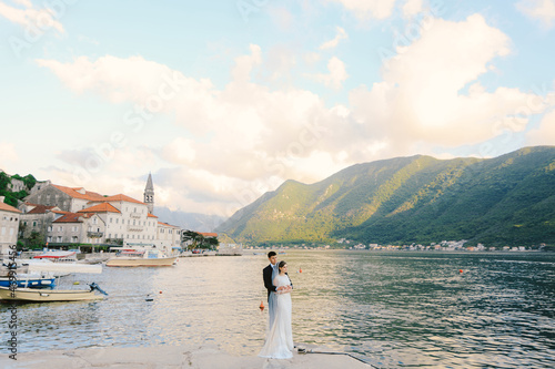 Groom hugs bride from behind while standing on the pier against the backdrop of the bay, mountains and ancient buildings