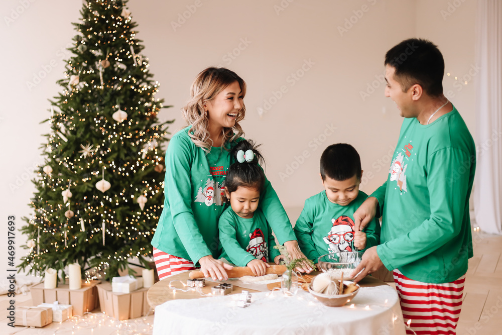 A happy mixed Asian family with two children in green pajamas cook cookies together and prepare for the Christmas holiday on New Year's weekend in a decorated room at home. Selective focus A happy mixed Asian family with two children in green pajamas cook cookies together and prepare for the Christmas holiday on New Year's weekend in a decorated room at home. Selective focus