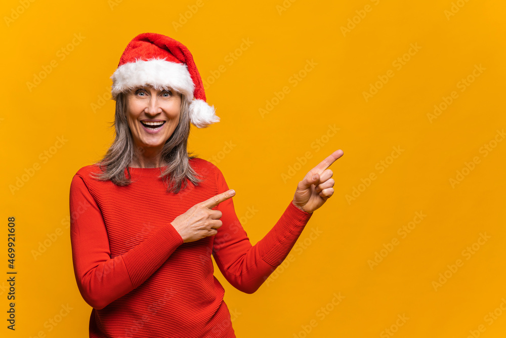 Fototapeta premium Excited senior grey-haired woman wearing red santa hat points fingers aside at empty copy space, presenting novelty, advertising product. Mature lady promoting new year deal, holiday sale