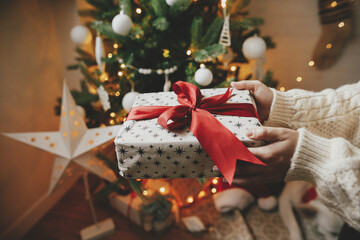Wrapped christmas gift with red bow in hands in cozy knitted sweater on background of christmas tree with lights. Stylish woman holding present in festive decorated room close up. Merry Christmas