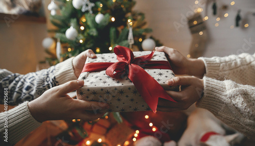 Fotografia Merry Christmas! Hands exchanging christmas gift with red bow on background of christmas tree with lights