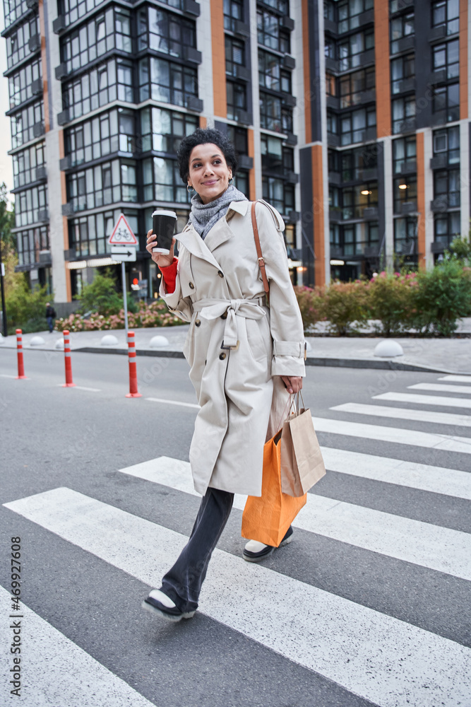 Calm multiracial woman walking with a paper cup of coffee and holding shopping bags Calm multiracial woman walking with a paper cup of coffee and holding shopping bags