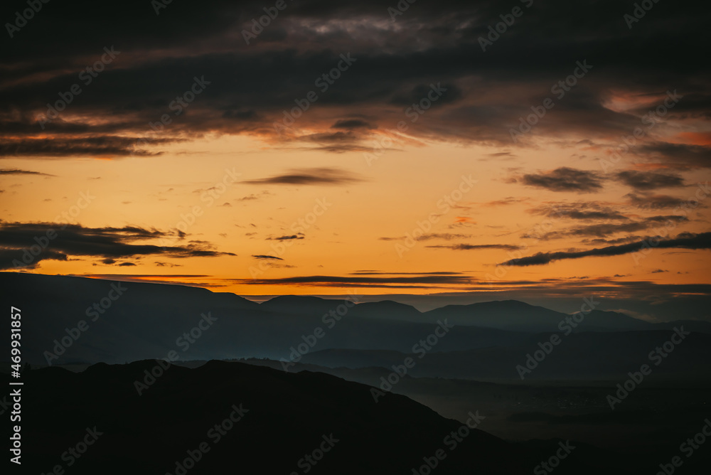 Scenic dawn mountain landscape with golden low clouds in valley among dark mountains silhouettes under sunset or sunrise sky. Vivid scenery with low clouds in mountain valley in illuminating color.
